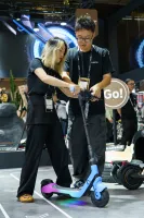 Zwei Personen testen zusammen einen blauen E-Scooter auf einer Messe.