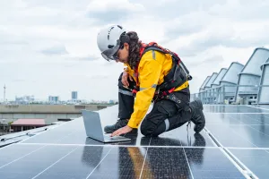Technikerin überprüft Solarpaneele auf einem Dach mit Laptop und Sicherheitsausrüstung.