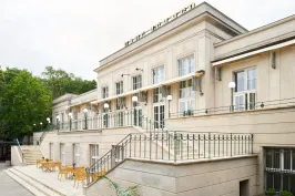 Bright building with outdoor terrace, tables and chairs, surrounded by trees.