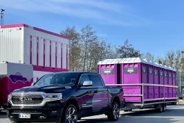 Hier wird ein Pick Up geuegt, der einen Anhänger zieht, auf dem mehrere, pinke Wölkchen WC-Kabinen stehen. Im Hintergrund ist ein WC-Wagen zu sehen, neben dem zwei, aufeinander stehende WC-Container, mit pinker Umrandung stehen.