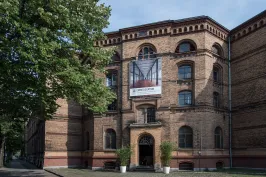 Backsteingebäude mit großem Werbebanner am Eingang, davor zwei Pflanzen.