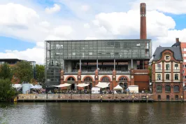 Backstein-Gebäude mit Glasfassade und Schornstein am Fluss, davor Menschen und Zelte.