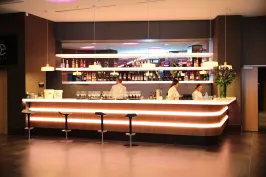 Moderne Bar mit drei Barhockern, zwei Barkeepern und beleuchteten Regalen im Hintergrund.
