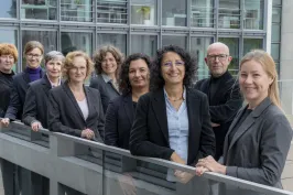 Neun Personen im Business-Outfit stehen lächelnd vor einem modernen Gebäude.
