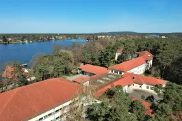 Gebäude mit roten Dächern am See, umgeben von Bäumen unter blauem Himmel.