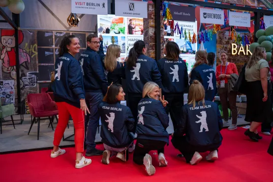 Gruppe von Frauen mit blauen Jacken und Bärenlogo posiert auf rotem Teppich bei einer Messe.
