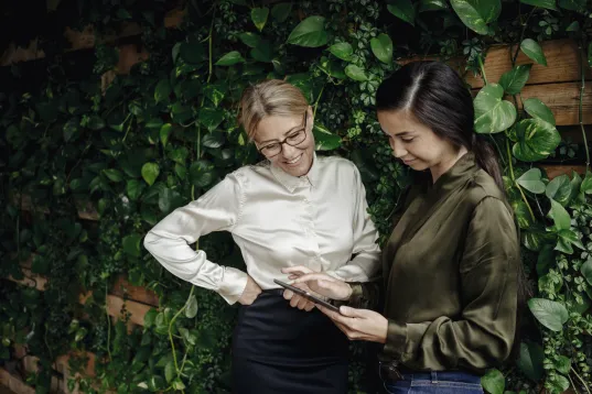 Zwei Frauen betrachten zusammen ein Tablet vor einer grünen Pflanzenwand.