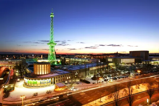 Beleuchteter Funkturm der Messe Berlin bei Abenddämmerung, umgeben von Gebäuden und Straßen.