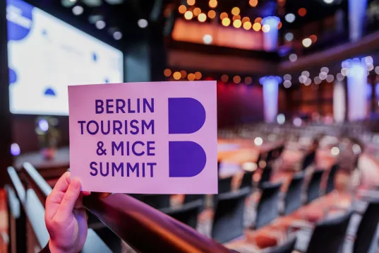 Hand hält Schild „Berlin Tourism & MICE Summit“ in leerem Konferenzsaal.
