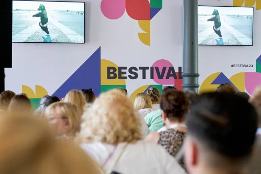 Publikum sitzt vor einer bunten Wand mit dem Schriftzug „Bestival“ und Monitoren.