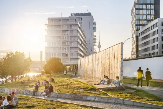 Menschen entspannen im Park an der Berliner Mauer bei Sonnenuntergang, moderne Gebäude im Hintergrund.
