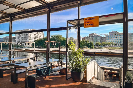 Überdachte Terrasse mit Sitzbänken, Flussblick und modernen Gebäuden im Hintergrund