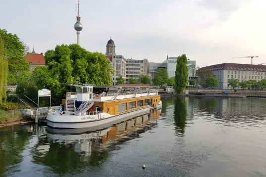 Ausflugsschiff auf einem Fluss in Berlin mit Fernsehturm und grünen Bäumen im Hintergrund.
