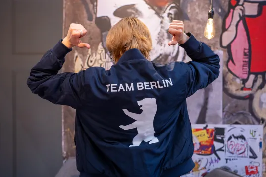 Person zeigt auf Jacke mit Bärenlogo und der Aufschrift "Team Berlin" vor Graffiti-Wand.