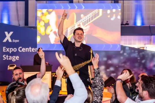 Ein Saxophonist in einer jubelnden Gruppe, im Hintergrund ist der Schriftzug "X Expo Expo Stage" zu erkennen