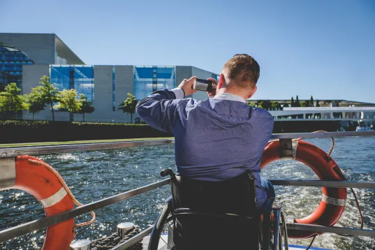 Auf dem Foto ist ein Mensch im Rollstuhl auf einer Schifffahrt zu sehen. 