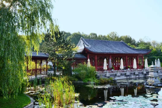 Auf dem Foto ist Roter asiatischer Pavillon mit Sonnenschirmen und grüner Umgebung bei klarem Himmel
