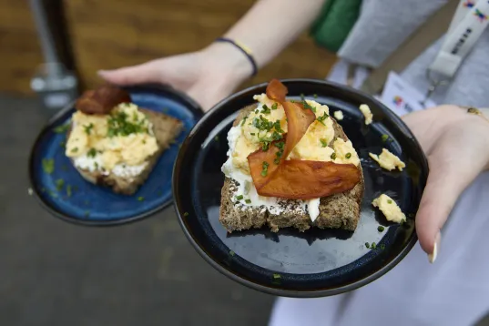 Person hält zwei Teller mit Brot, Rührei, Frischkäse, Schnittlauch und gebackenem Gemüsechips.