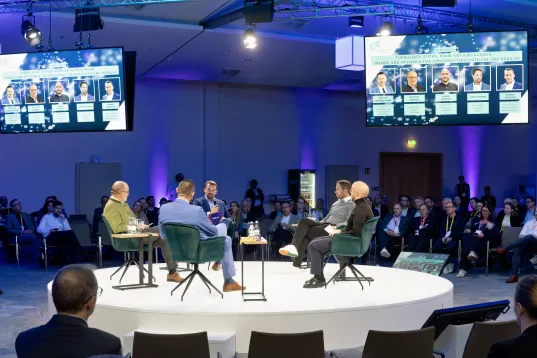 Eine runde Plattform, auf der Personen miteinander diskutieren und Teilnehmende, die um die Bühne herum sitzen.