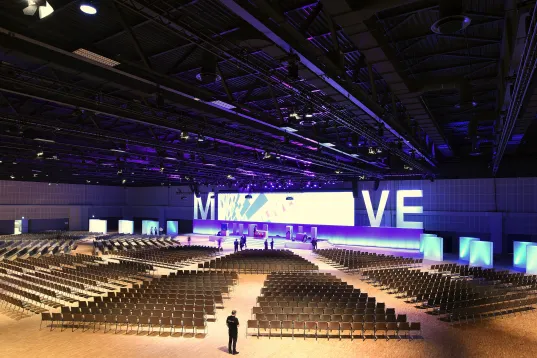 Großer, moderner Konferenzsaal mit Bühne, vielen Stuhlreihen und lila Beleuchtung.