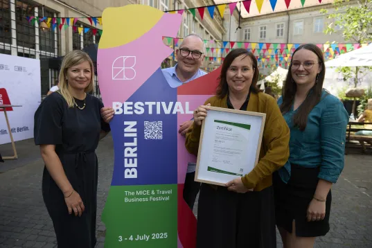 Vier lächelnde Personen posieren draußen mit einer Urkunde vor einem bunten Festival-Schild.