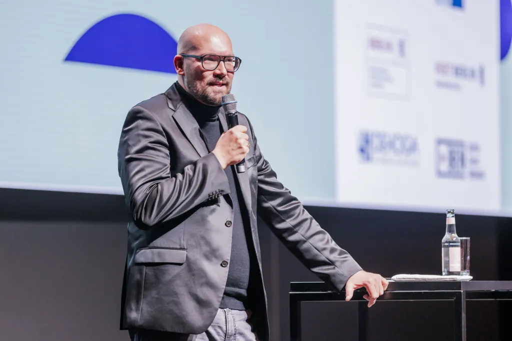 Staatssekretär Michael Biel spricht auf Bühne in Mikro mit Präsentation im Hintergrund