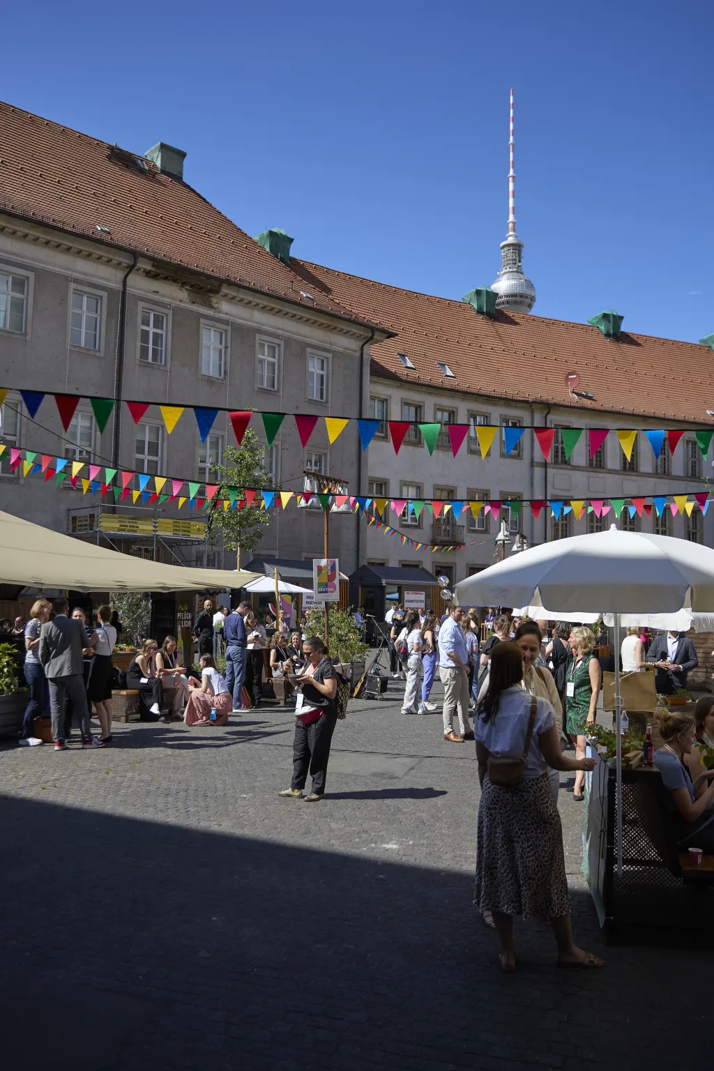 Belebter Innenhof der Alten Münze mit Wimpeln, Marktständen und Menschen unter Sonnenschirmen bei blauem Himmel.
